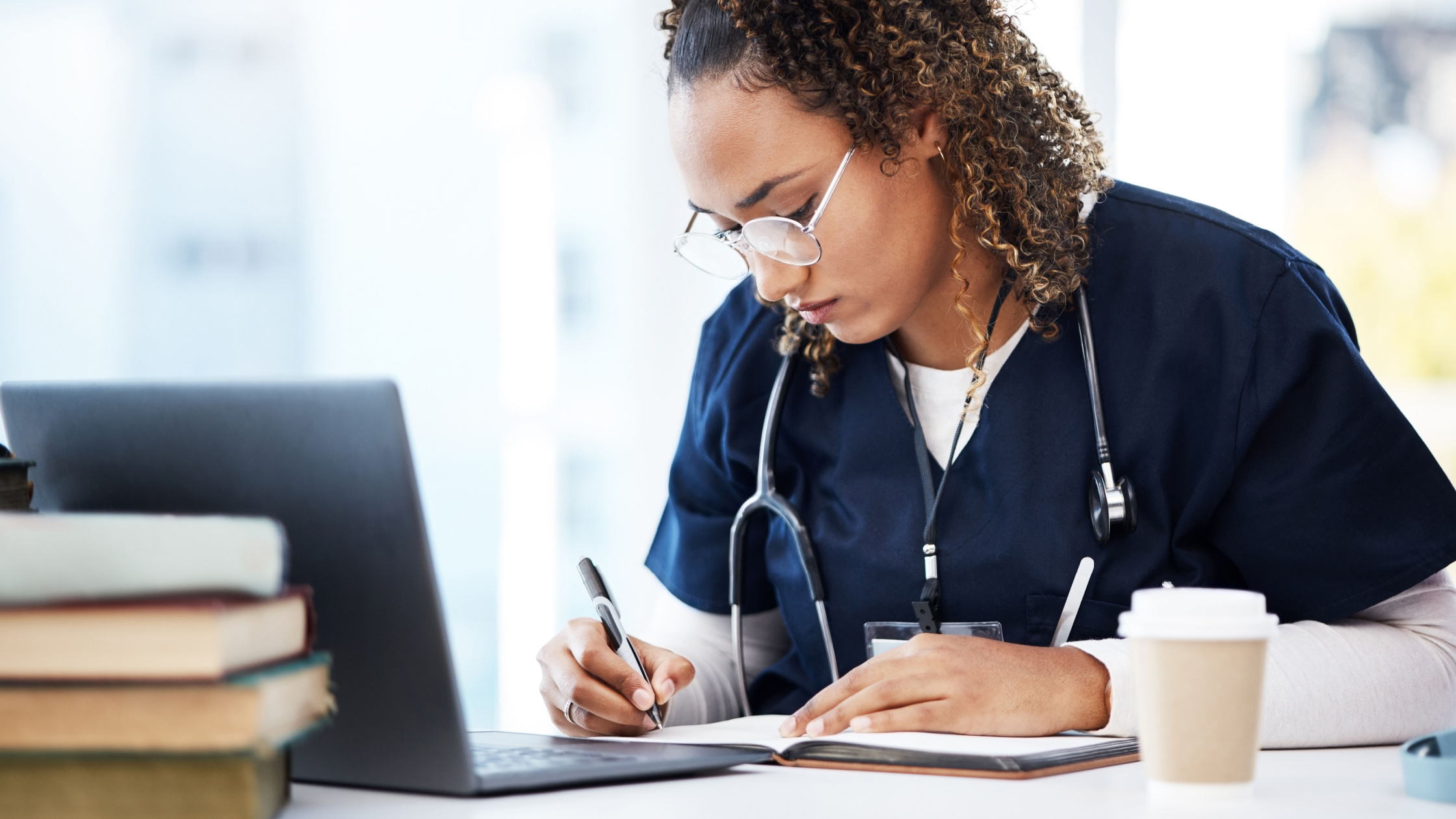 A medical student studying for her psychiatry shelf exam, wearing scrubs, in front of a laptop and textbooks.