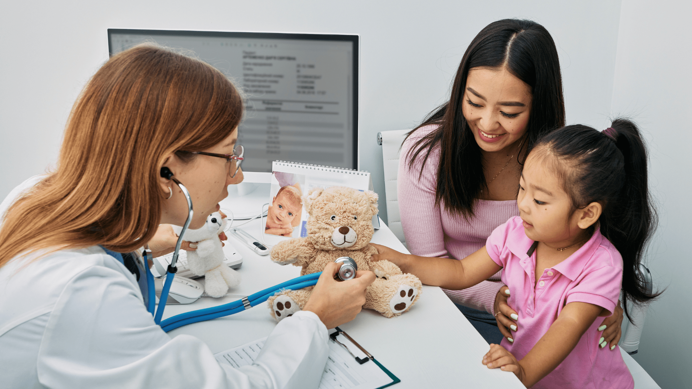 Female doctor using a stethoscope on a teddy bear to engage a young girl and her mother during a pediatric check-up in a medical office.