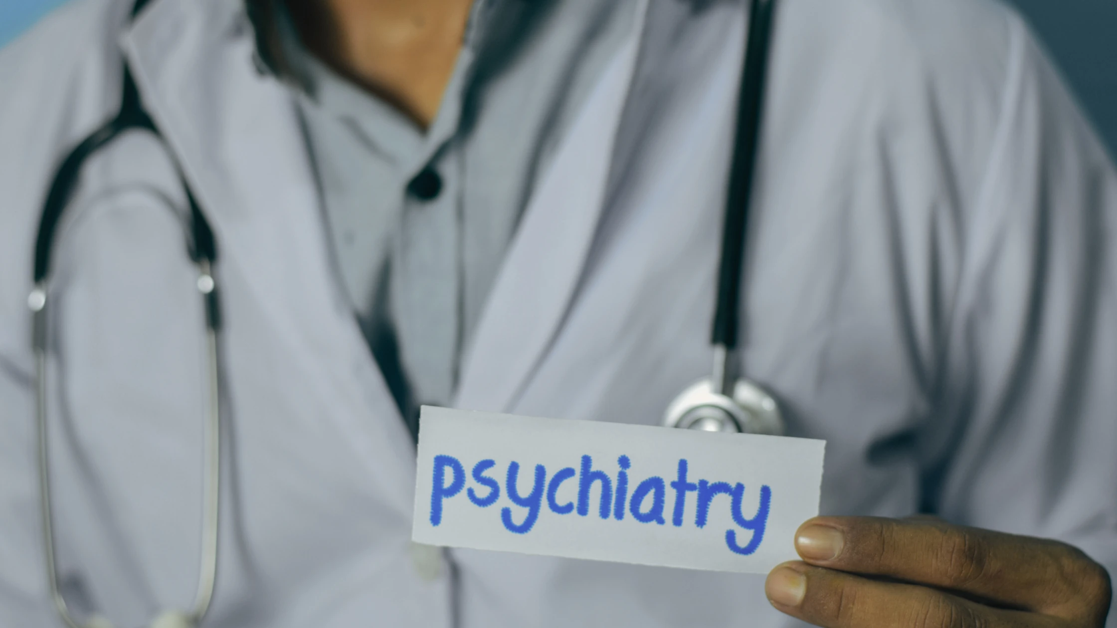 A doctor in a white coat, holding a card with the word "psychiatry" written on it, symbolizing the focus of the blog on achieving a high score in the Psychiatry Shelf Exam.