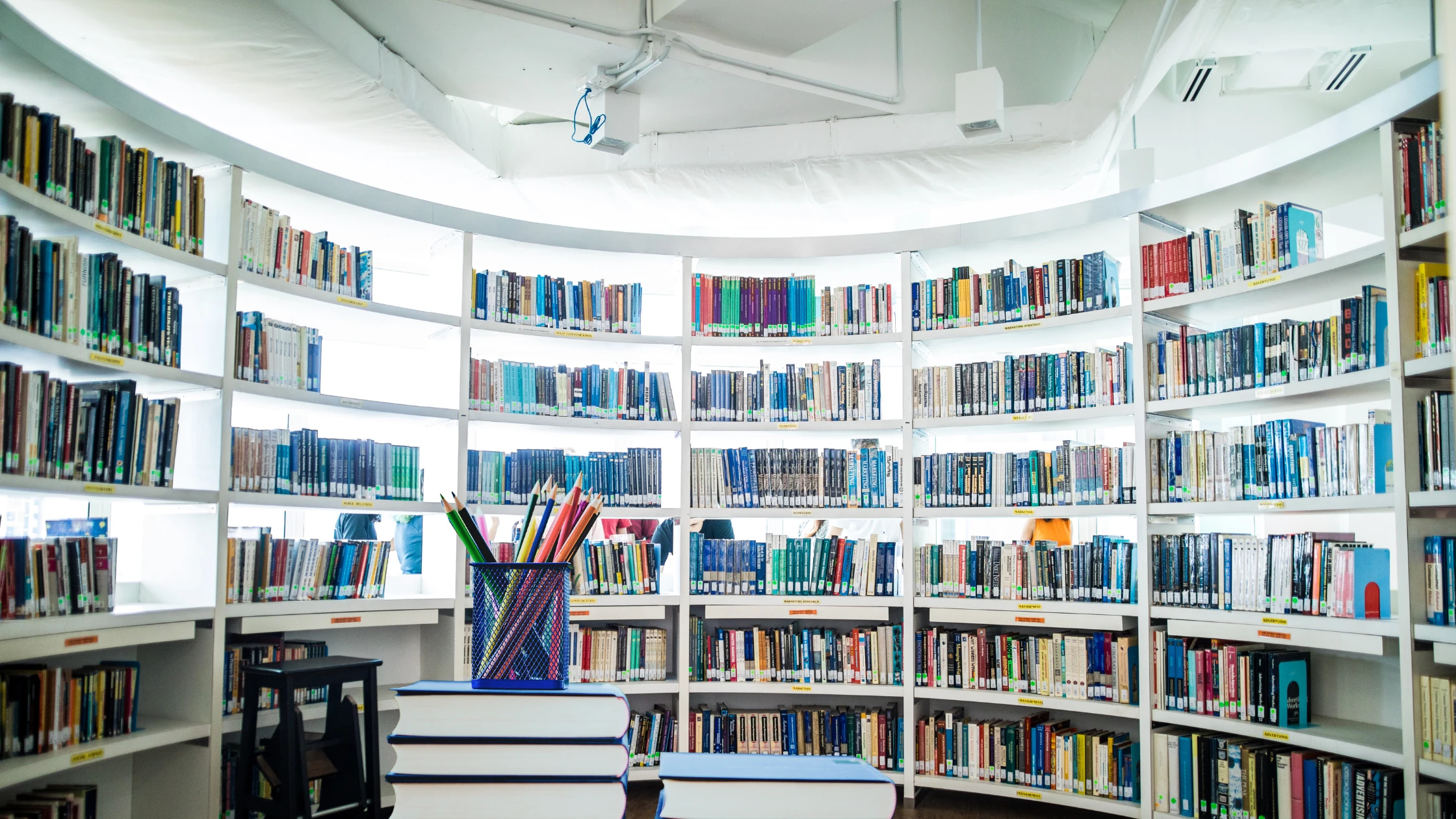 Circular library with white shelves full of books and a pencil holder on stacked books in the foreground.