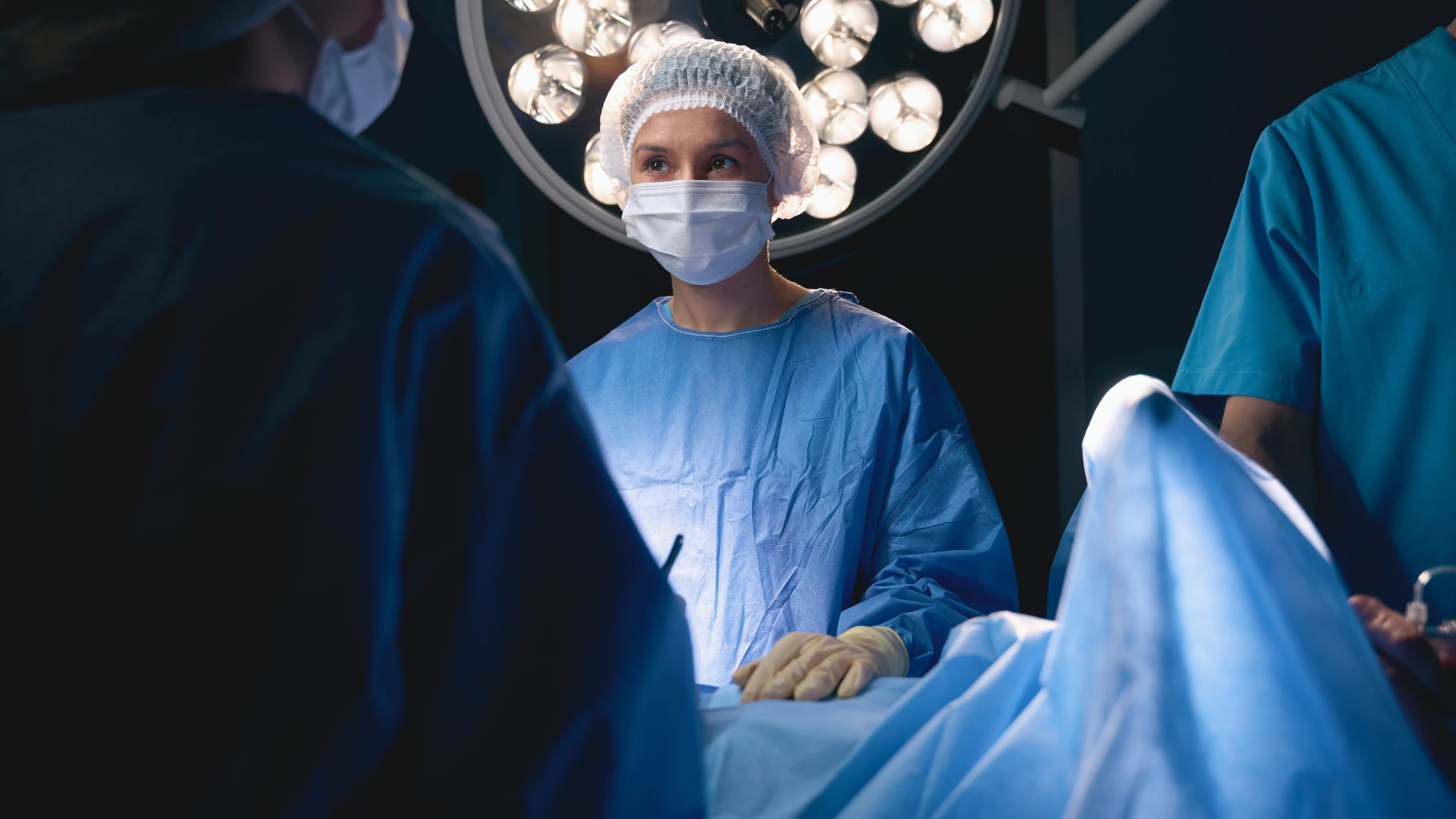 Female surgeon in scrubs and mask standing in an operating room, illuminated by surgical lights overhead.