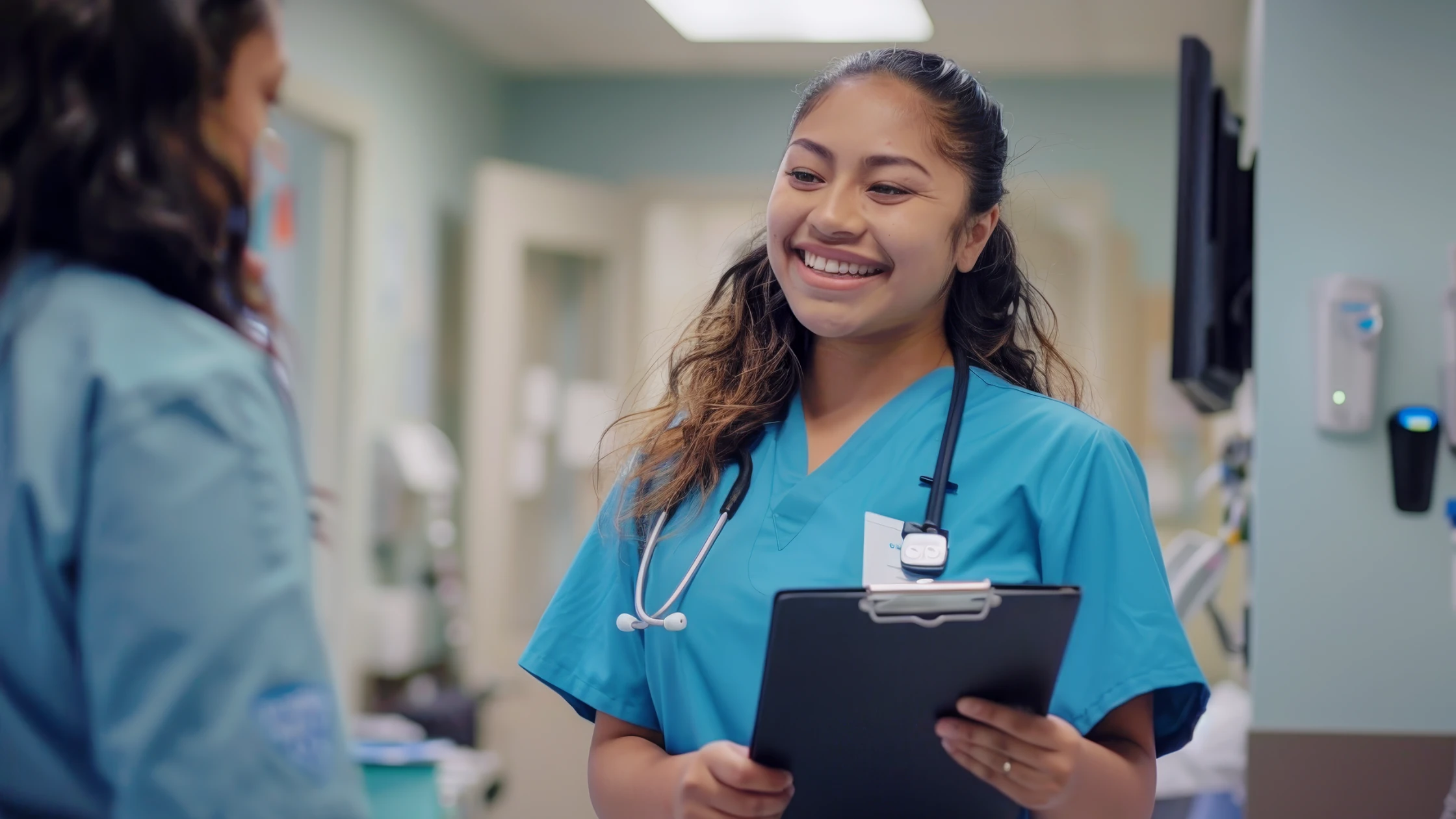 Medical student in blue scrubs holding a clipboard, engaged in a conversation in a healthcare volunteering environment.